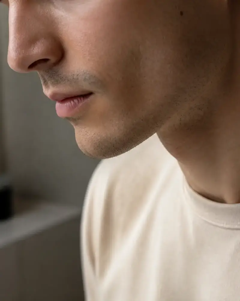 Profil d’un visage d’homme penché légèrement vers le bas, éclairage doux de fenêtre, ligne de la mâchoire visible, personne non identifiable