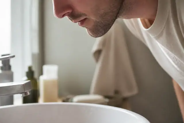Menton et joue d’un homme penché sur un lavabo clair, lumière douce de fenêtre, peau légèrement rosée, personne non identifiable